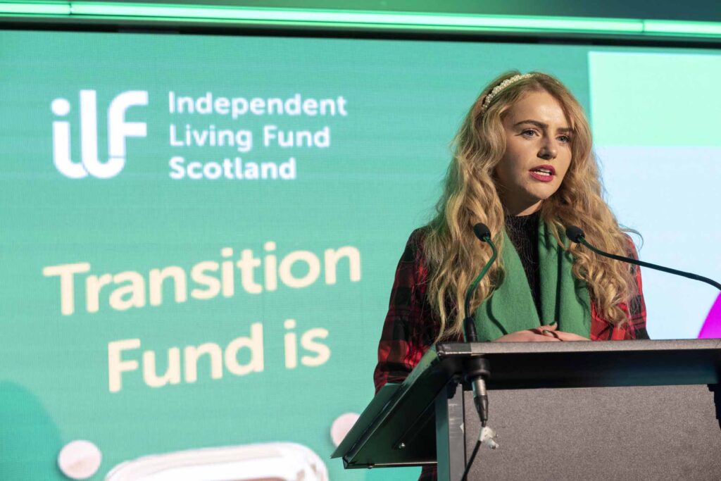 Young woman with long blonde hair standing in front of a podium. In the background is the ILF Scotland logo and the words "Transition Fund".