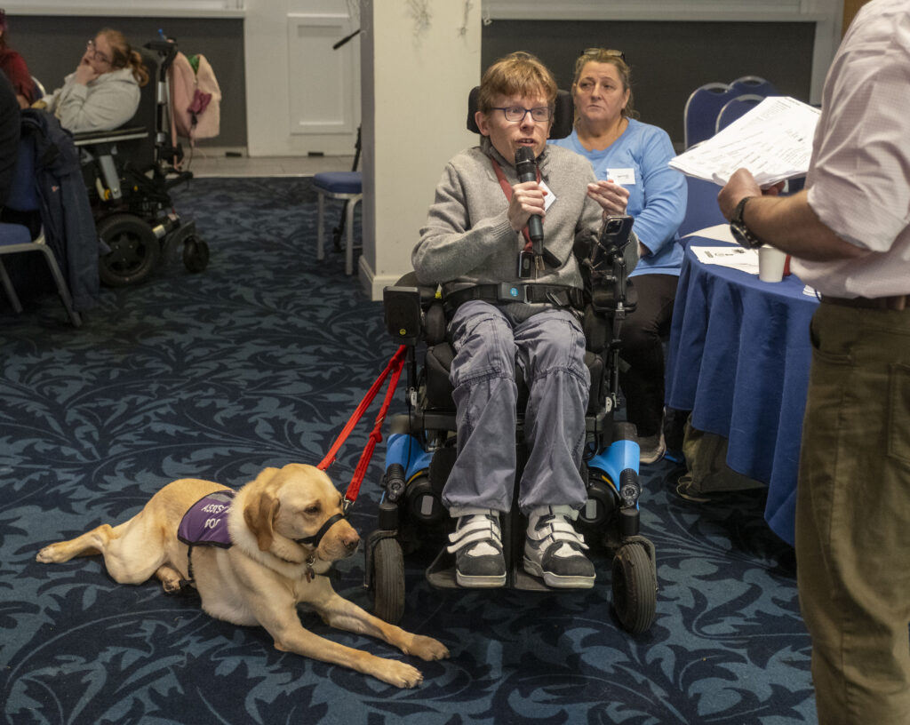 A person addresses the room with a microphone. A guide dog lies beside them.