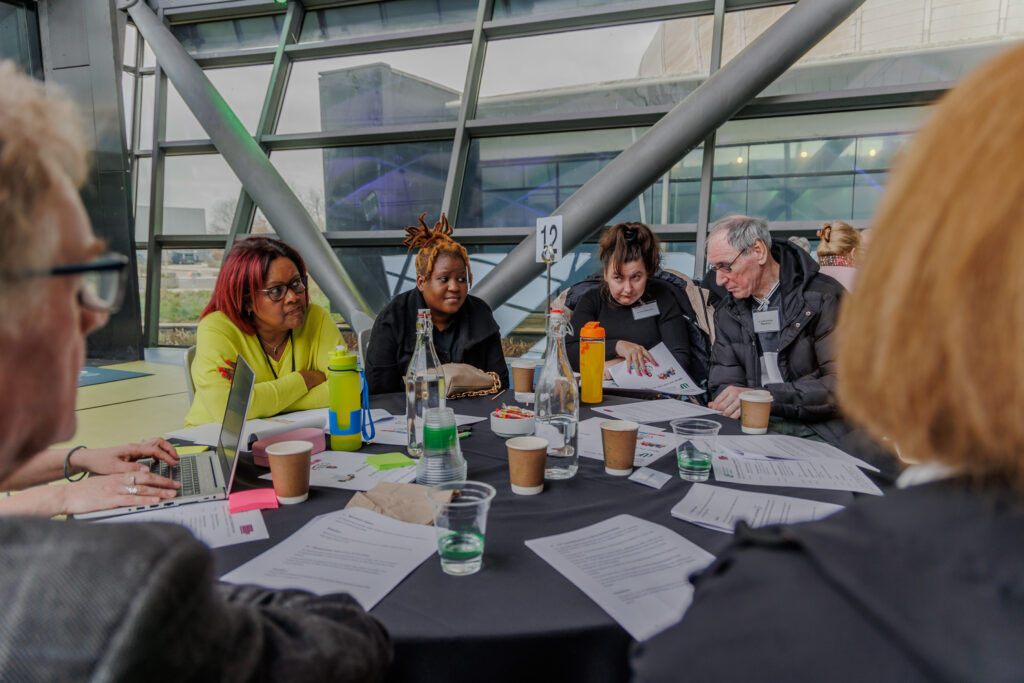 A photo of attendees of ILF Scotland's co-production event at Glasgow Science Centre. A group of people are seated at a table covered with documents.
