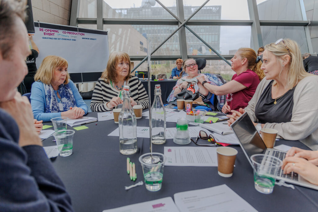 A photo of attendees of ILF Scotland's co-production event at Glasgow Science Centre. A group of people are seated at a table covered with documents. There is a presentation board in the background with the words 'Re-Opening Co-Production Event'.