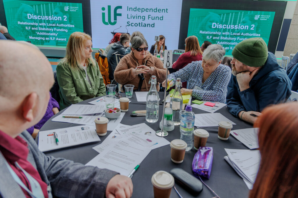A photo of attendees of ILF Scotland's co-production event at Glasgow Science Centre. A group of people are seated at a table that is covered in documents. Behind them are three display screens with the Independent Living Fund Scotland logo and the words 'Discussion 2'.