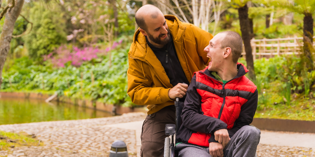 Two men are at a park. One man leans down behind the other who is in a wheelchair. They are looking at each other and smiling and laughing. Behind them you see the flowers, a bridge and a pond in the park.