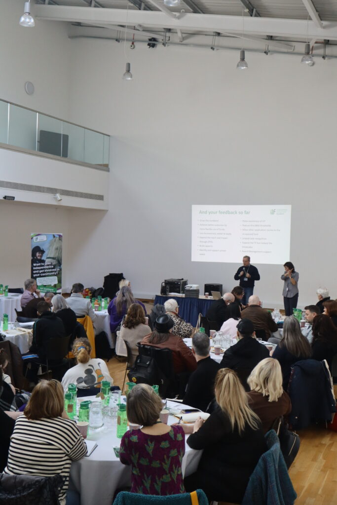 A view of two people at the front of a hall with a white screen behind them. Tables of people at round tables watch what's happening.