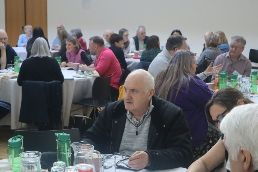 A view of tables of people chatting and gathering in a hall. At the forefront of the picture is a man looking to someone off camera.