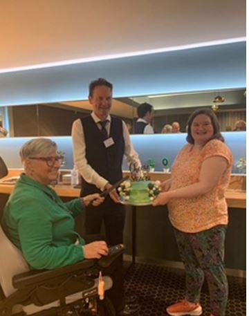 Two people holding a green cake between them. A lady in a wheelchair holds a knife to cut the cake.