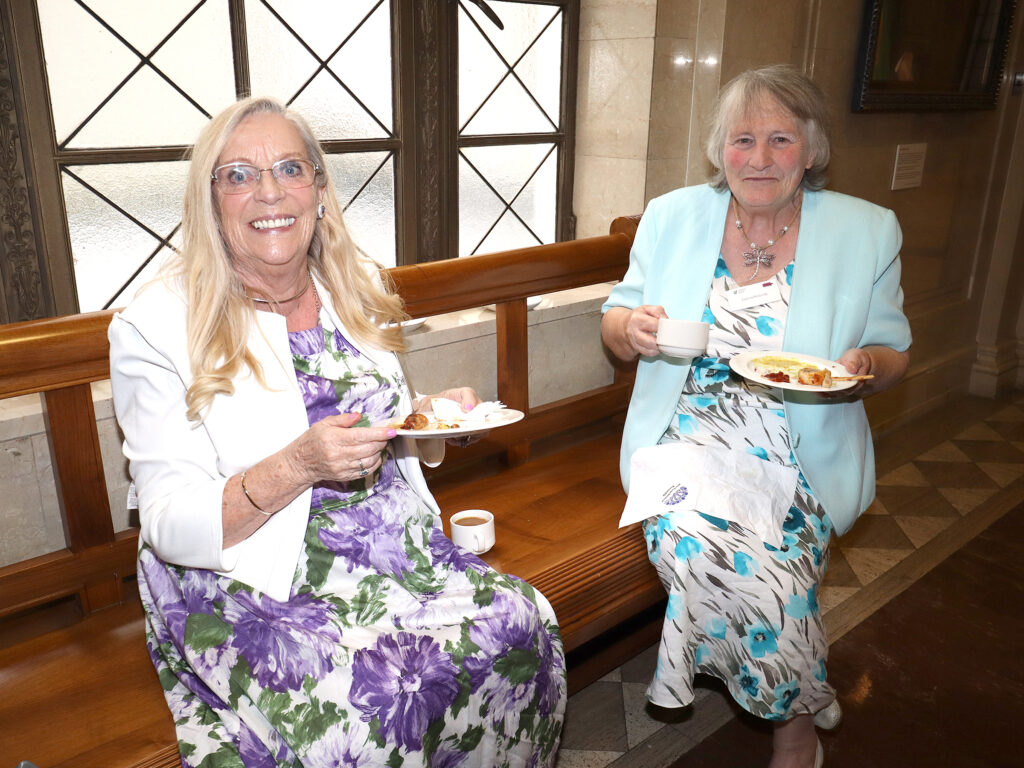 Two ladies in floral dresses sit at a window, with plates.