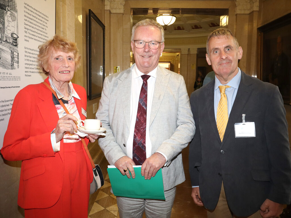 A lady with a tea cup stands beside two men - all posing for the camera.