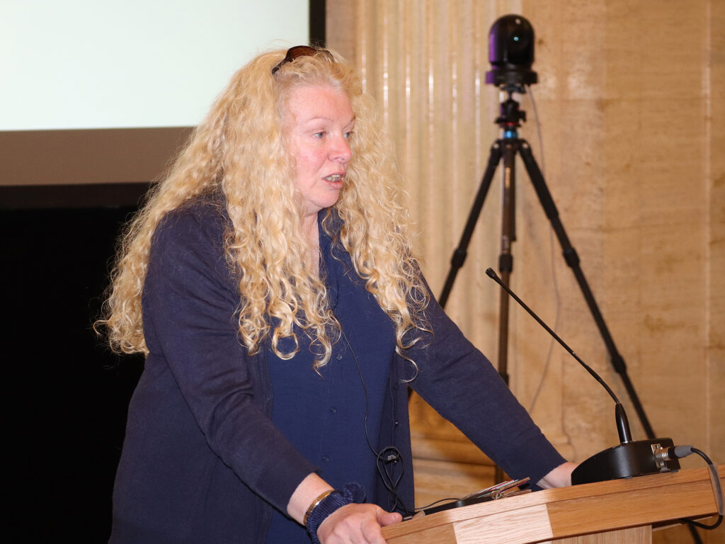A lady with blonde, curly hair stands at the podium.