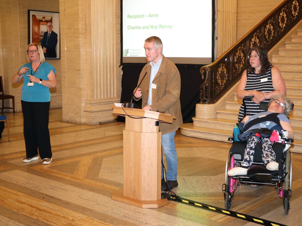 A man stands at the podium. His wife and sister are off to one side. The ISL interpreter stands on the other.