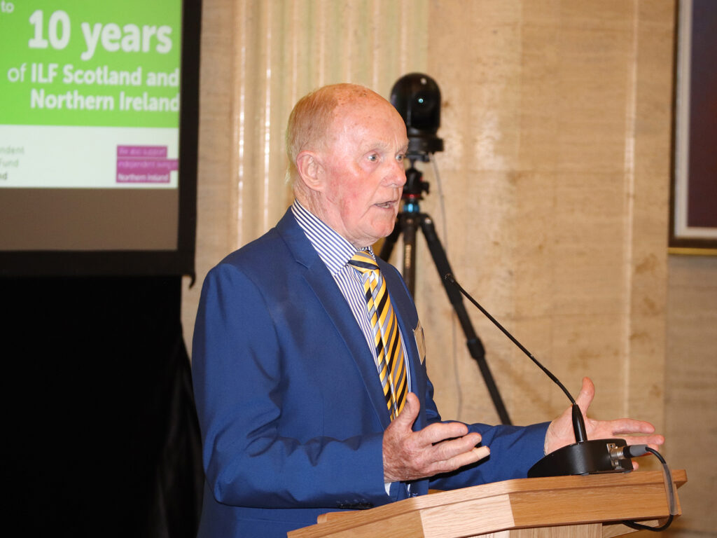 A former MLA stands at the podium and speaks to the audience.