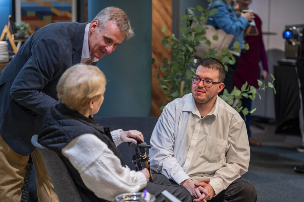 A man leans over to speak to two people who are sitting down.