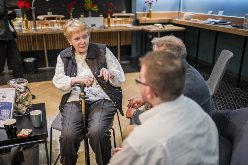 A lady sits in a chair talking to two men, who we can see the backs of their heads.