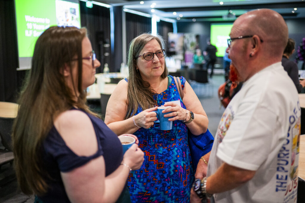 Two ladies, both drinking coffee, speak to a man in a white shirt.