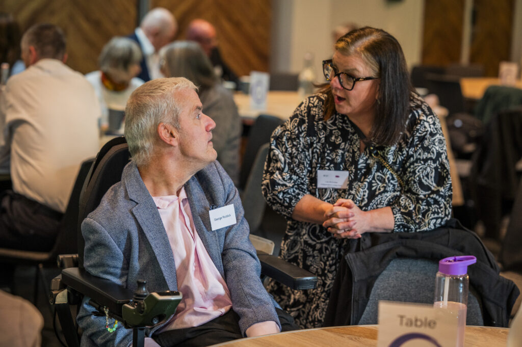 A lady standing and talking to a man seated at a table.