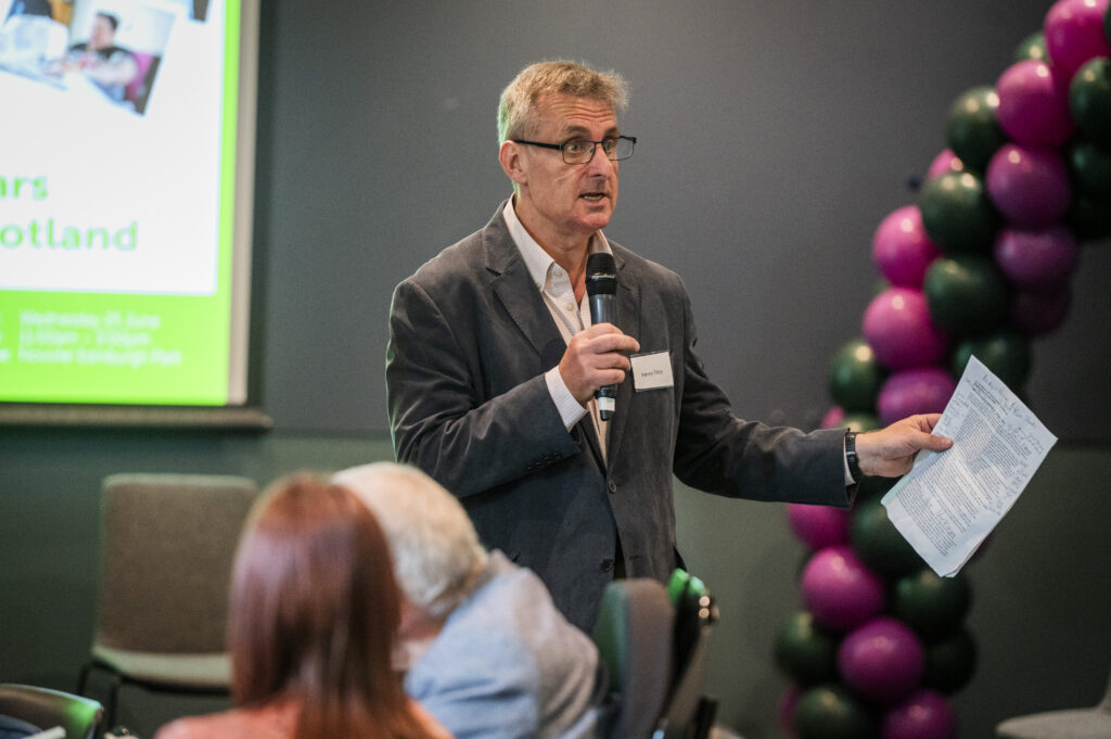 Harvey standing, speaking with a microphone.