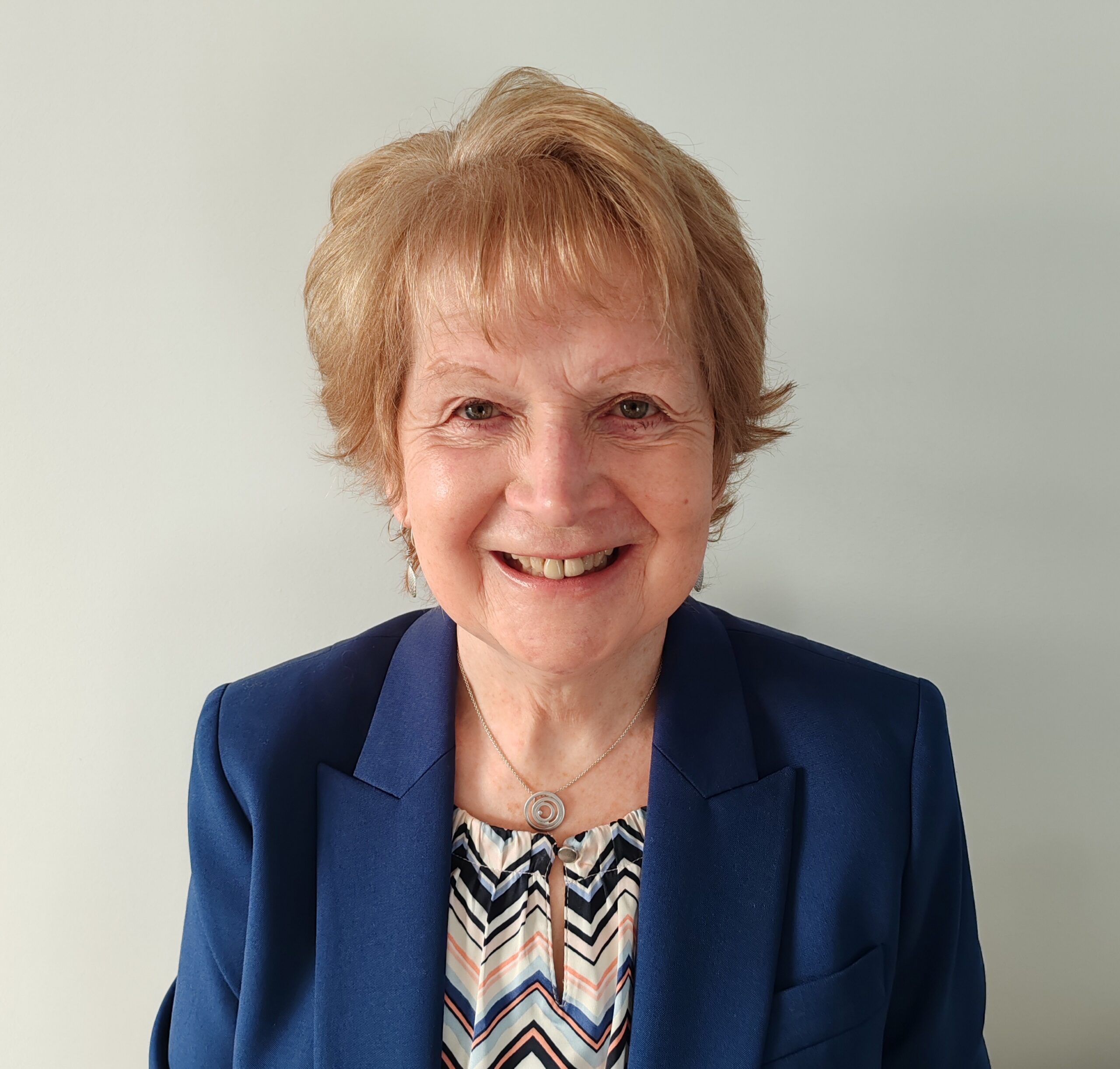 Photograph of a lady with blond hair wearing a patterned top and blue jacket. She's smiling