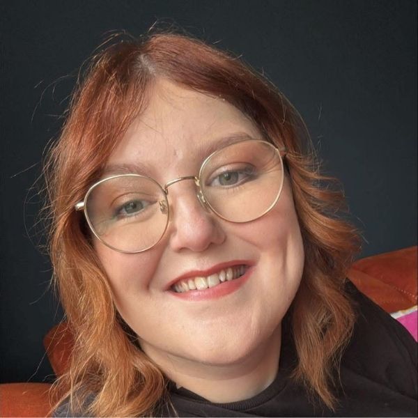 Dan has short curly red hair and glasses with a thin gold rim. She is also wearing red lipstick and a black top. 