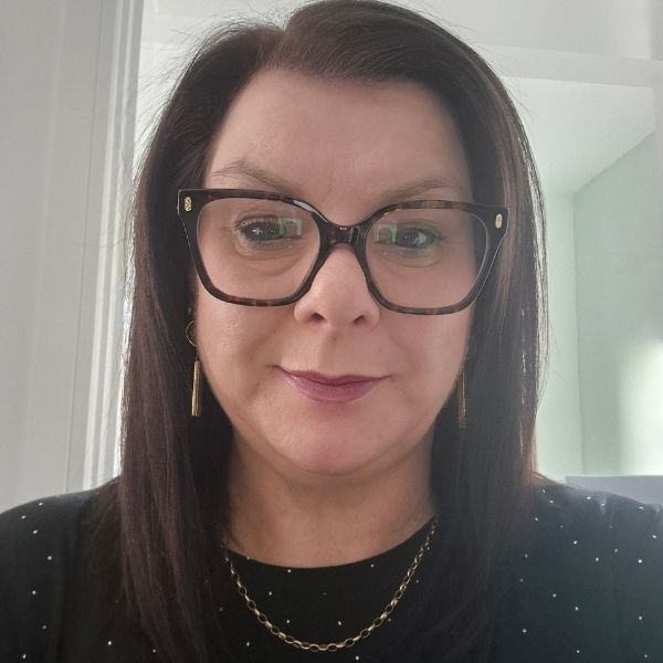 Head and shoulders photo of Lisa, who has short dark brown hair and is wearing dark framed glasses, a black jumper with white polka dots and a gold chain necklace.