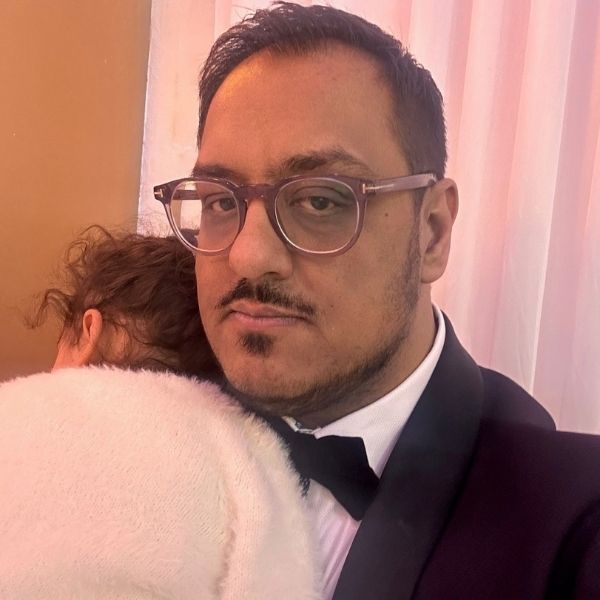 Photo of Nadeem who has short black hair and is wearing a black suit with black bowtie, and purple coloured glasses. He is holding his daughter so that her face is not seen and she is wearing a white fluffy jumper. 