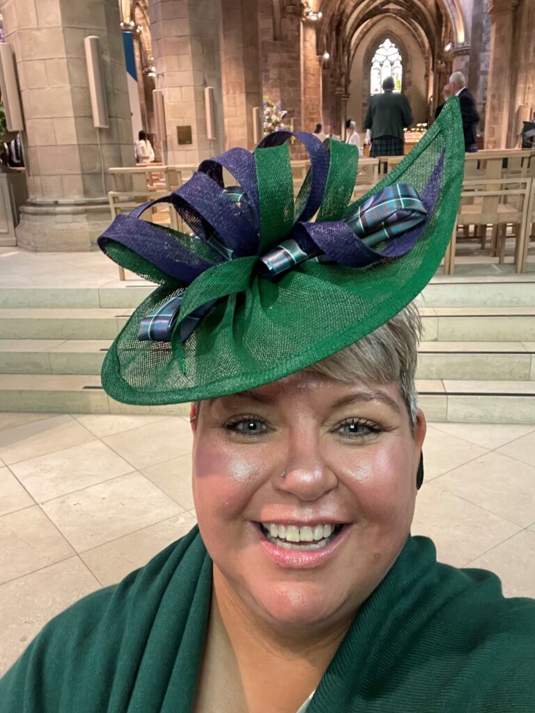Heather smiling, wearing a green hat decorated with green, purple, and tartan ribbons, and a green top.
